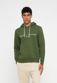 Zöld kapucnis pulóver kenguruzsebbel; fehér "Tommy Hilfiger" logóval és alapítási dátummal, zsinóros kapucnival és bordás mandzsettákkal.