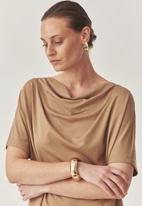 Beige draperad topp i mjukt tyg, guld böjd armband och guld triangulära örhängen kompletterar den mjuka looken. Enkel, modern design.