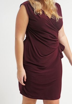 Vrouw draagt een mouwloze, bordeauxrode wikkeljurk met gedrapeerde stofdetails aan de voorkant tegen een effen achtergrond.