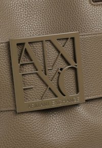 Borsa in pelle marrone texturizzata caratterizzata da un logo metallico geometrico con la scritta "Armani Exchange". Il logo è ben visibile e progettato con linee nette.