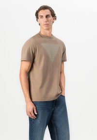 Joven de pie con la mano en el bolsillo, vistiendo una camiseta marrón con un sutil diseño triangular y jeans azules, contra un fondo blanco.