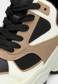 Χοντρό αθλητικό παπούτσι με τμήματα από μαύρο, μπεζ και λευκό δέρμα, μαύρα κορδόνια, υφή στη σόλα και στρωματοποιημένες σχεδιαστικές λεπτομέρειες.