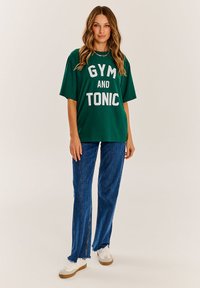 Zelené nadrozmerné tričko s nápisom "GYM AND TONIC" vytlačeným na bielo, skombinované s modrými denimovými nohavicami s rozšírenými nohavicami a bielymi teniskami so svetlo hnedými podrážkami.