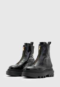 Botines de piel negra con un acabado brillante, que cuentan con una cremallera dorada en la parte frontal, suela de goma negra gruesa y una lengüeta en la parte posterior.