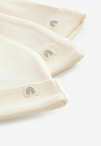 Hellbeige gestrickte Mützen mit umgeschlagenen Bündchen, versehen mit kleinen rechteckigen Etiketten, die mit einem Regenbogenlogo in gedämpftem Grauton geprägt sind.