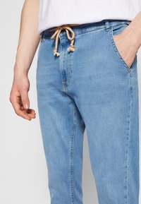 Svetlo modri jeans hlače z vrvico na pasu, ki imajo stranske žepe in gladko, teksturirano površino. Očitni šivi za poudarjanje.