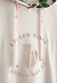 Beiger Hoodie aus strukturiertem Stoff, mit einem erhöhten "b"-Logo und dem Schriftzug "by Ted Baker". Verziert mit einer Kapuze mit floralem Muster und Kordeln.