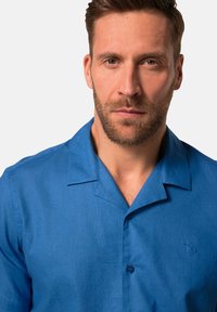 Camicia azzurra a maniche corte con bottoni, realizzata in tessuto intrecciato con una texture liscia, accentuata da un piccolo logo ricamato sul petto sinistro.