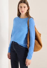 Hellblauer Pullover mit gerippten Bündchen, kombiniert mit dunklen Jeans. Eine braune Wildleder-Umhängetasche hängt von einer Schulter. Minimalistischer Hintergrund.
