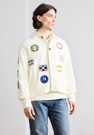 Young man with shoulder-length dark hair wearing a cream jacket with colorful patches and blue jeans, standing against a plain background.