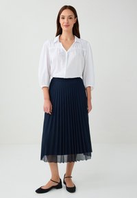 Camicia bianca con dettagli arricciati, abbinata a una gonna midi plissettata di un blu scuro e ballerine nere con cinturini alla caviglia.