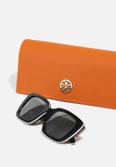 Svarta fyrkantiga solglasögon med mörka linser, placerade bredvid ett orange hårt fodral med en guldlogotyp. Båda föremålen ligger platt på en vit bakgrund.