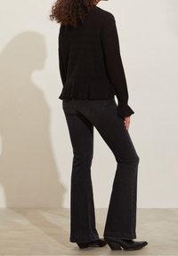 Pull en maille texturée noire avec un ourlet et des manchons à volants. Associé à un jean évasé noir et des bottes à talons noires. Fond simple et lisse.