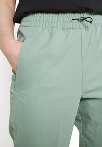 Person med ljusgröna byxor med elastisk midja och svart snörning, hand placerad i vänster ficka.