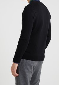 Black sweater with a crew neck, fitted design, ribbed cuffs and hem. Worn over grey trousers, showing a woven blue and green check pattern underneath.