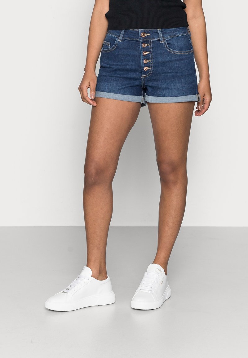 Person som har på sig rullade mörkblå denimshorts med fem framskjutna knappar och vita sneakers, stående på ett grått golv.
