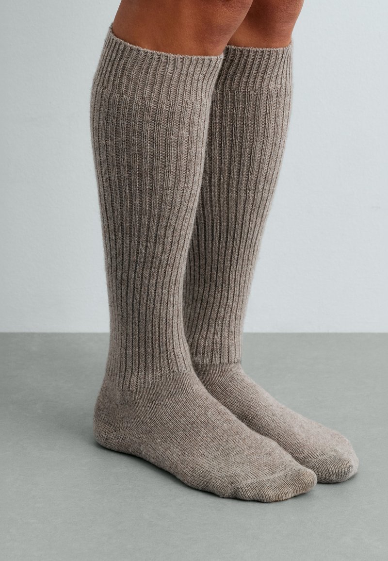 Jambes portant des chaussettes en laine côtelées montant jusqu'aux genoux, de couleur beige neutre, debout sur une surface grise avec un fond clair uni.