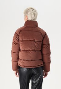 Brun cordfløyel pufferjakke med høy krage og horisontal quiltet design. Kombinert med svarte faux skinnbukser som viser en teksturert finish.