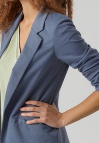 Světle modrý blazer s texturovanou tkaninou, přiléhavým střihem a otevřeným límcem. Obsahuje jedno přední kapsu a ohrnuté rukávy.