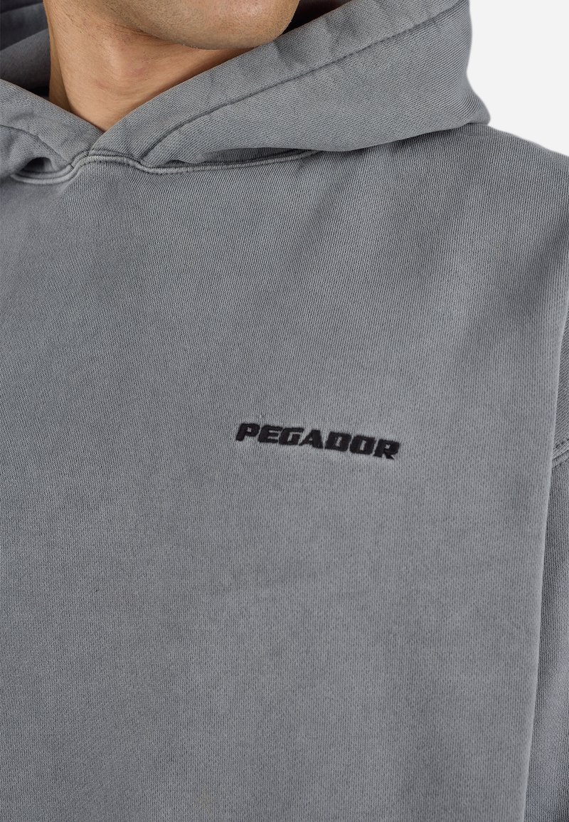 Sudadera gris claro con capucha ajustable, con el logo bordado en negro "PEGADOR" en el frente. Tejido suave y ligeramente texturizado.