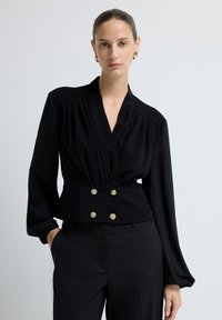 Sort bluse med lange ærmer, dyb V-hals, plisseret design, indsnævret talje og knapdetaljer i guldtoner; parret med skræddersyede sorte bukser.
