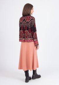 Vrouw staat rechtop, gekleed in een zwart en rood gebloemd jack, lange perzikkleurige rok en zwarte enkellaarsjes, met haar zijwaarts gericht tegen een witte achtergrond.