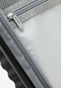 Primer plano de una cremallera gris con "WITTCHEN" en la lengüeta del tirador y gotas de agua sobre una superficie lisa de color gris claro debajo.