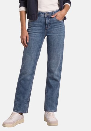 Person iført blå jeans med lige ben, hvide sneakers, en hvid skjorte og en mørk jakke med den ene hånd i lommen og den anden afslappet.