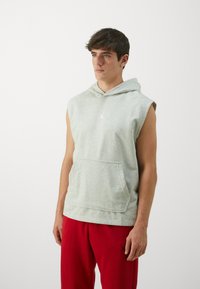 Grå ærmeløs hoodie med en frontlomme, der har et hvidt logo på brystet. Bæres med røde bukser og har en blød tekstur.