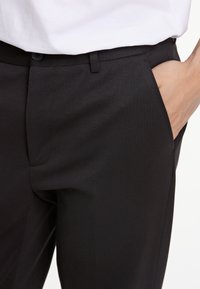 Schwarze, maßgeschneiderte Hose mit glatter Textur und dezentem Glanz. Verfügt über Seitentaschen und einen eleganten Bund mit Knopfverschluss.