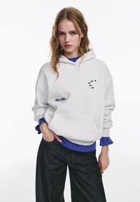 Witte hoodie met zak aan de voorkant en een zwarte sterrenprint. Gelaagd met een blauw topje, gedragen met donker wijde jeans. Zacht materiaal, ontspannen pasvorm.