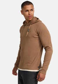 Felpa con cappuccio lunga a maniche, color marrone con un tascone a kangaroo sul davanti e un orlo bianco a contrasto. Presenta coulisse e un'etichetta con il logo sul petto.