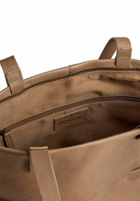 Interno di una borsa in pelle marrone con una tasca con cerniera e etichetta con il logo. Texture morbida, design aperto e manici doppi visibili.