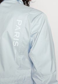 Ljusblå jacka i slätt material, med vit text "PARIS" och "SAINT-GERMAIN" tryckt vertikalt på sidan.