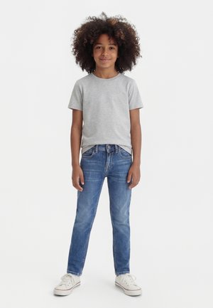 Bambino con capelli ricci in piedi che indossa una t-shirt grigio chiaro, jeans blu e scarpe da ginnastica bianche contro uno sfondo bianco uniforme.