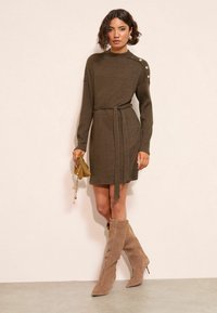 Abito a maniche lunghe in maglia a coste marrone con cintura da annodare. Presenta dettagli con bottoni su una spalla. Abbinato a stivali con tacco color cammello.