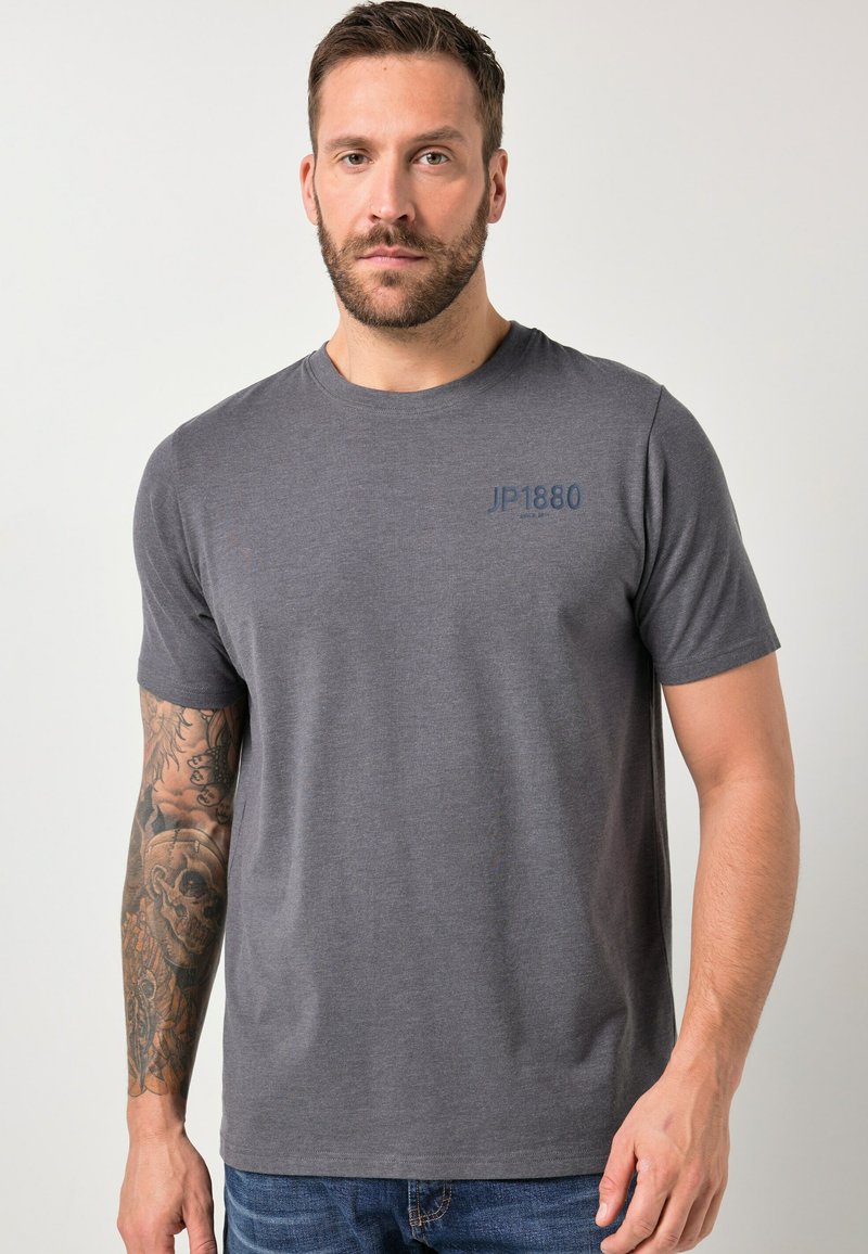 Homme aux cheveux bruns courts et barbe, portant un T-shirt gris avec le texte "JP1880" et un jean bleu, debout devant un fond clair uni.