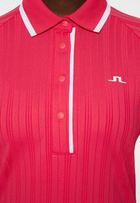 Czerwona koszulka polo z pionowym prążkowanym wzorem, białymi akcentami na kołnierzyku, pięcioma dopasowanymi guzikami oraz małym logo nadrukowanym na biało.