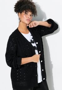 Černý pletený kardigan s jemným otevřeným vzorem, s véčkovým výstřihem, dlouhými rukávy a bílými knoflíkovými detaily. Stylizovaný přes bílé tričko.