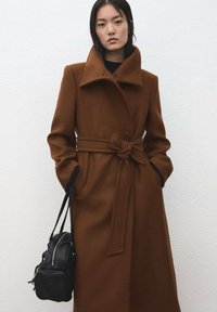 Bruine wollen jas met een hoge kraag, tailleband en klepzakken. Heeft een gladde textuur en een zwarte accenten op de mouwen.