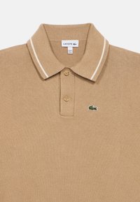 Beżowa bawełniana koszulka polo z klasycznym kołnierzykiem, dwoma guzikami i białymi paskami. Na piersi znajduje się małe zielone logo Lacoste.