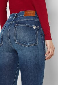Jeans de mezclilla azul oscuro con cintura de altura media, dos bolsillos traseros y una etiqueta de cuero marrón en la cinturilla; presenta una pequeña etiqueta blanca en el bolsillo.