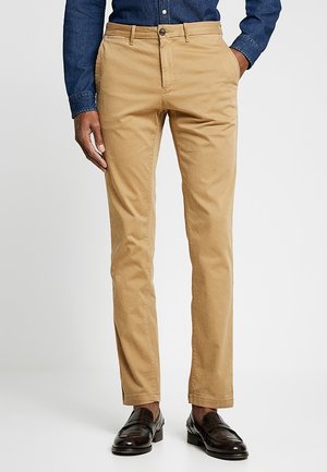 Homme portant un chino slim beige, des mocassins en cuir marron foncé et une chemise bleue à manches longues boutonnée, debout avec une main dans la poche.