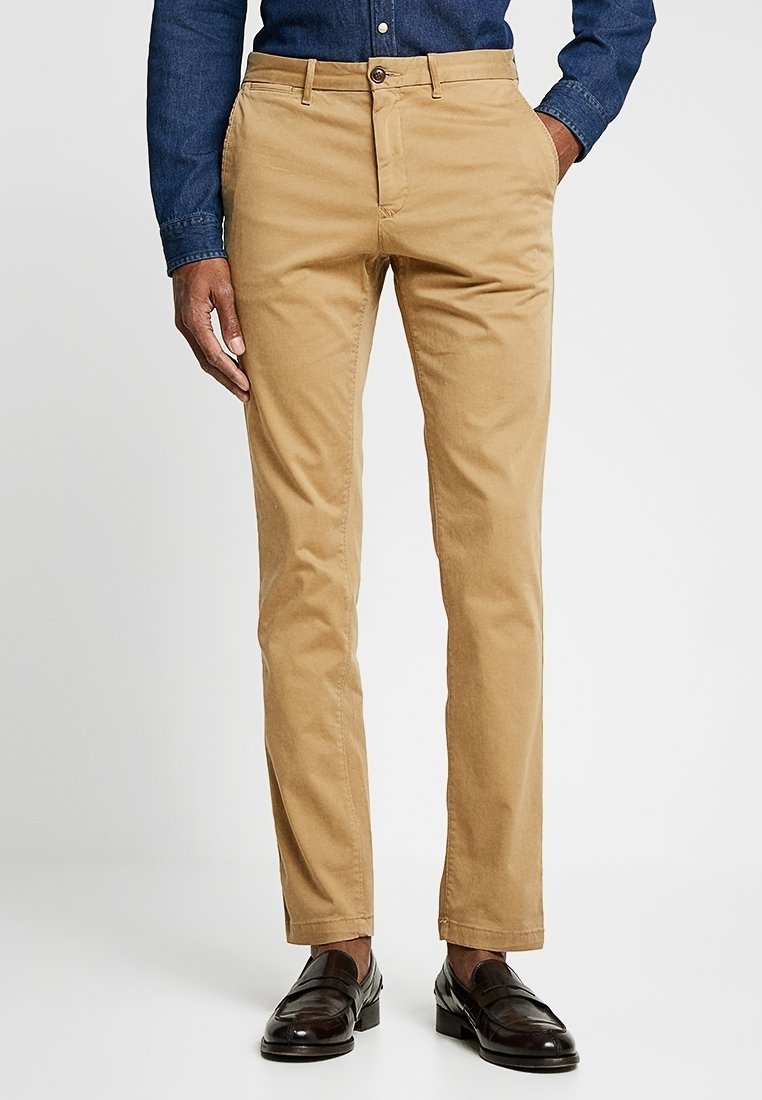 Homme portant un chino slim beige, des mocassins en cuir marron foncé et une chemise bleue à manches longues boutonnée, debout avec une main dans la poche.