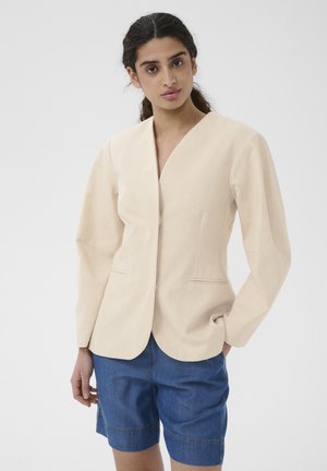 Žena s tmavými vlasmi, ktorá má na sebe béžové sako a modré rifle šortky, pózuje s jednou rukou v vrecku pred jednoduchým pozadím.