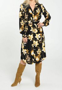 Robe noire à fleurs avec de grandes fleurs blanches et jaunes, manches longues, taille cintrée et longueur au genou. Associée à des bottes marron en suede à hauteur de genou.