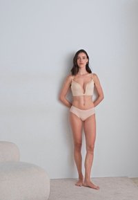 Nude spalvos liemenėlė be siūlių su gilia iškirpte ir reguliuojamomis petnešėlėmis, derinama su atitinkamomis žemomis kelnaitėmis. Minkšta medžiaga, minimalus dekoravimas.