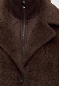 Gros plan d'un manteau marron duveteux avec un large col porté par-dessus une veste matelassée foncée zippée jusqu'au cou.