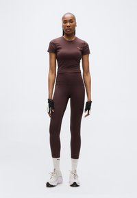 Αθλητική γυναίκα που στέκεται, φοράει σκούρο καφέ εφαρμοστό cropped τοπ και κολάν, λευκές κάλτσες, λευκά αθλητικά παπούτσια και μαύρα γάντια χωρίς δάχτυλα.