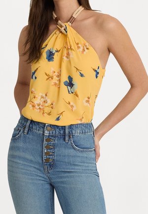 Kobieta w żółtym, kwiecistym topie na szyję z zaplecionym paskiem oraz w wysokich niebieskich dżinsach z pięcioma widocznymi guzikami.